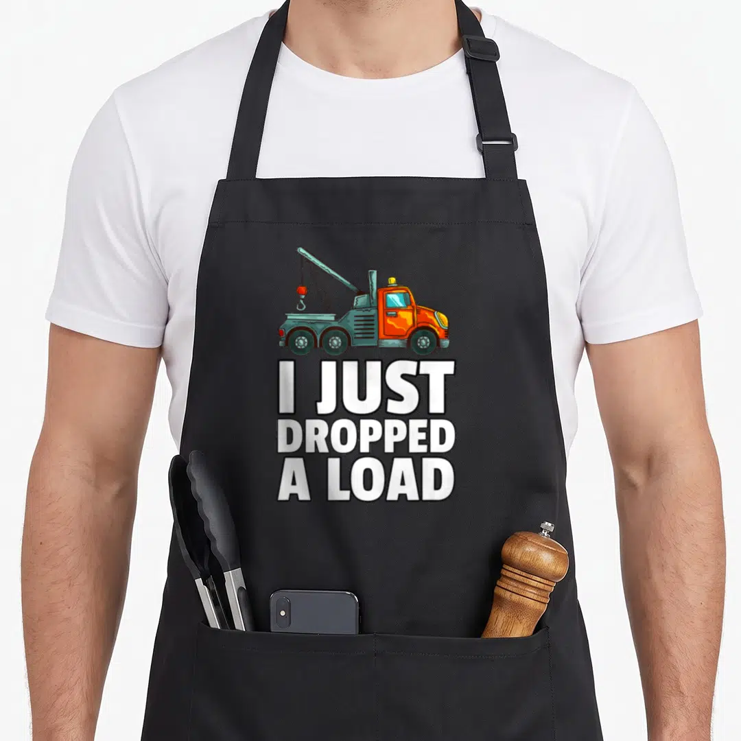 Driver operator truck apron - Cartoon orange and grey tow truck above stacked white text I JUST DROPPED A LOAD — shown on a black cotton canvas apron against a white background