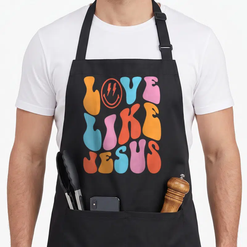 Jesus face on apron - Three stacked bubbly letters LOVE LIKE JESUS in orange red blue pink with red smiley face as O — shown on a black cotton canvas apron against a white background
