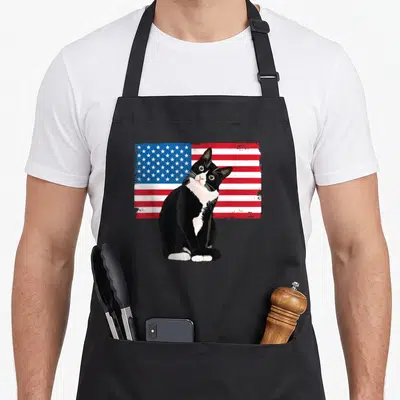 4th tuxedo apron - Realistic black and white cat sitting in front of a distressed USA flag background — shown on a black cotton canvas apron against a white background