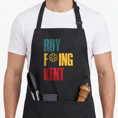 Freaking rob roy apron - Stacked ROY FOING KENT text in teal yellow red with geometric star symbol — shown on a black cotton canvas apron against a white background