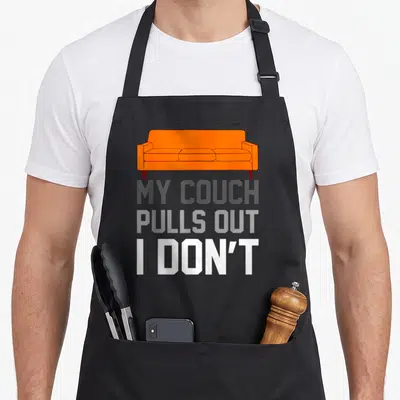 Couch offensive aprons - Orange cartoon pull-out sofa above stacked text MY COUCH PULLS OUT I DON'T in grey and white — shown on a black cotton canvas apron against a white background
