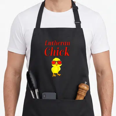 Reformation rose apron - Red text Lutheran Chick above yellow cartoon chick wearing red sunglasses — shown on a black cotton canvas apron against a white background