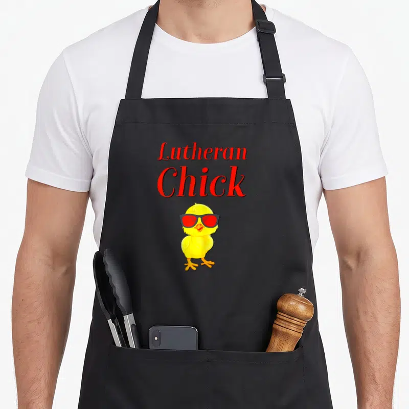 Reformation rose apron - Red text Lutheran Chick above yellow cartoon chick wearing red sunglasses — shown on a black cotton canvas apron against a white background