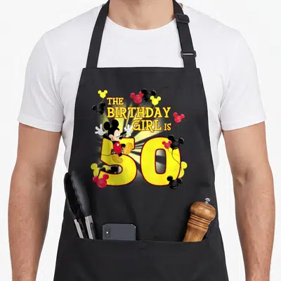 50th friends apron - Mickey Mouse sits on yellow 50 with red and black balloons and stacked text reading The Birthday — shown on a black cotton canvas apron against a white background