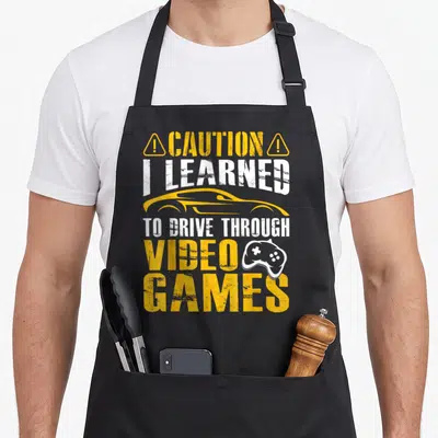 Permit driver wife apron - CAUTION I LEARNED TO DRIVE THROUGH VIDEO GAMES text with yellow car and white controller icon — shown on a black cotton canvas apron against a white background