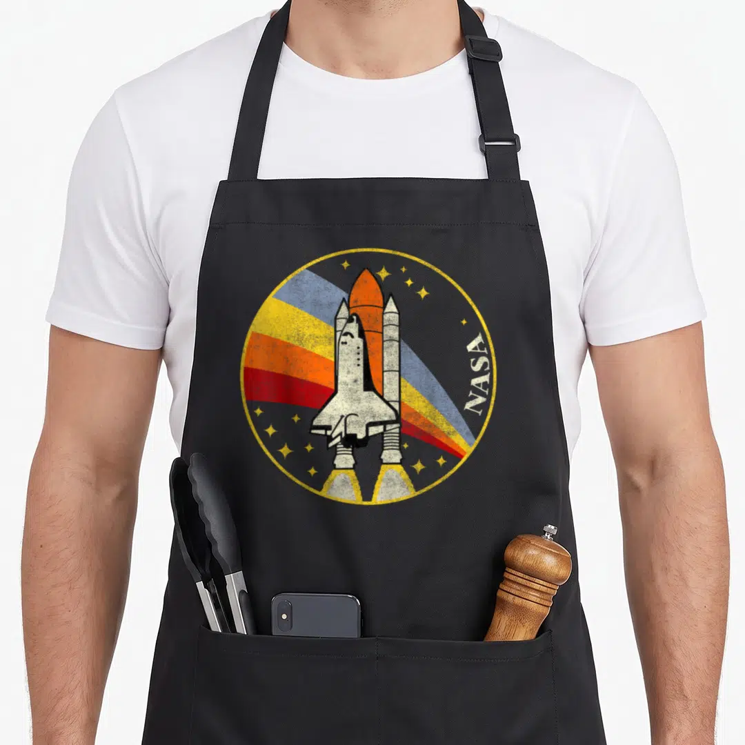 Distressed nasa apron - Retro space shuttle badge with rainbow stripes and vertical NASA text inside yellow circle — shown on a black cotton canvas apron against a white background