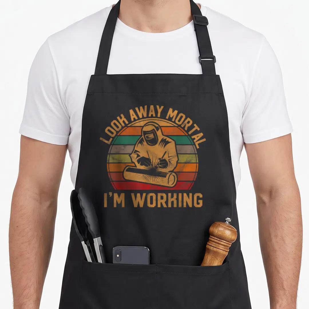 Away blacksmith apron - Retro welder silhouette with striped sunburst and text LOOK AWAY MORTAL I'M WORKING — shown on a black cotton canvas apron against a white background