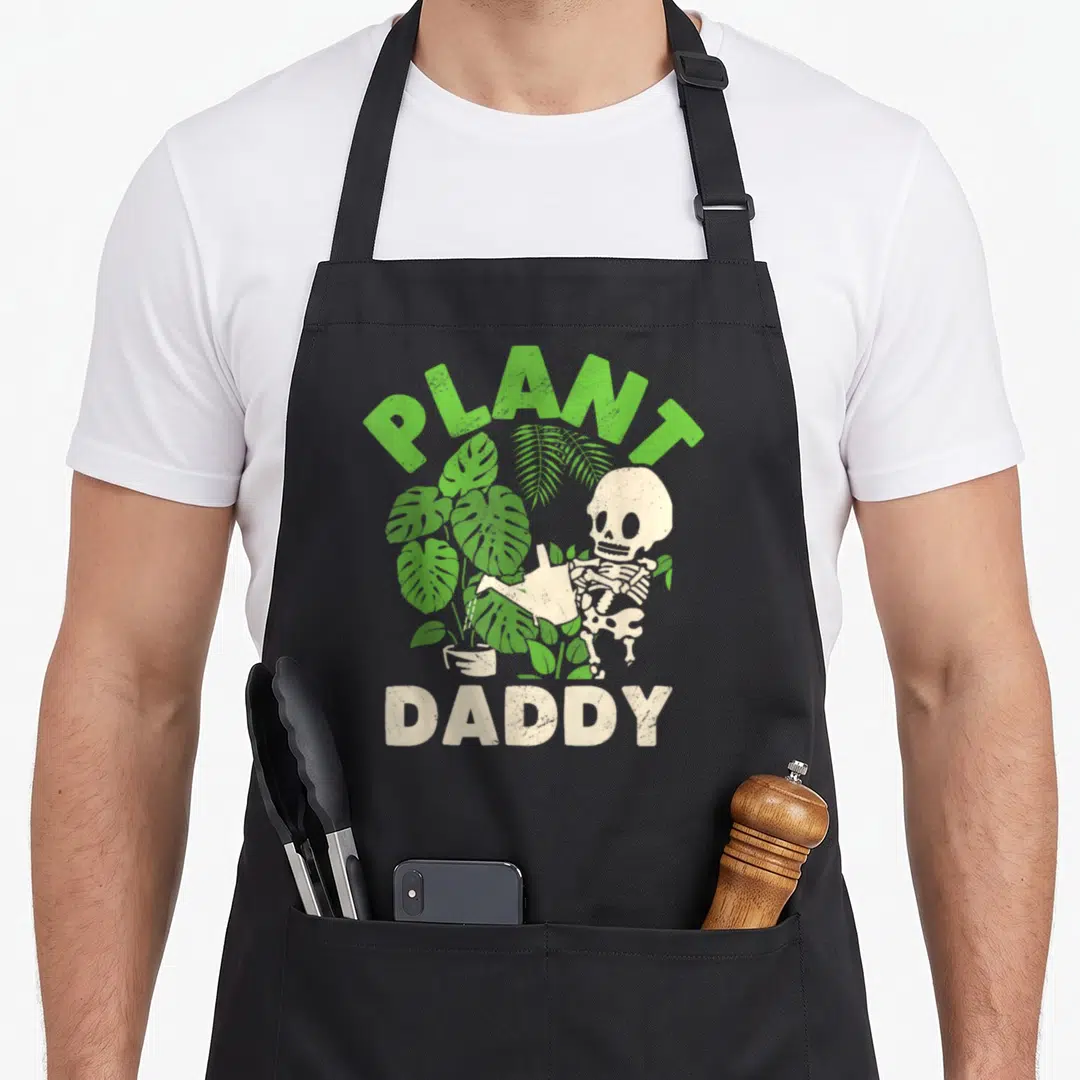 Daddy gardener apron - Skeleton watering green plants with PLANT DADDY text in green and beige — shown on a black cotton canvas apron against a white background