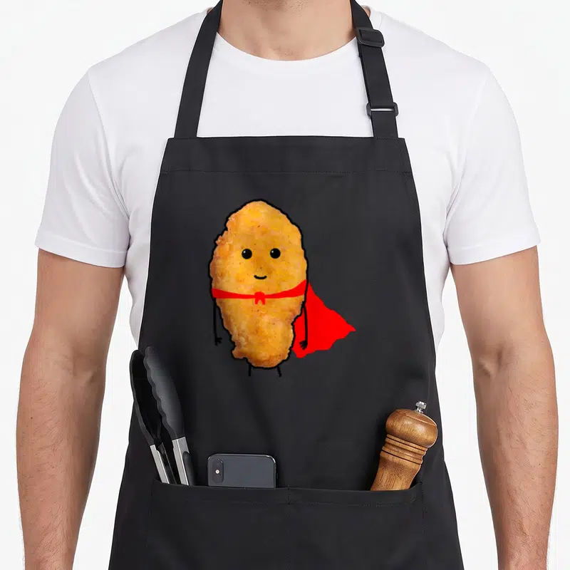 Hero chicken print apron - Cartoon golden brown chicken nugget wearing red cape with black limbs and smile — shown on a black cotton canvas apron against a white background