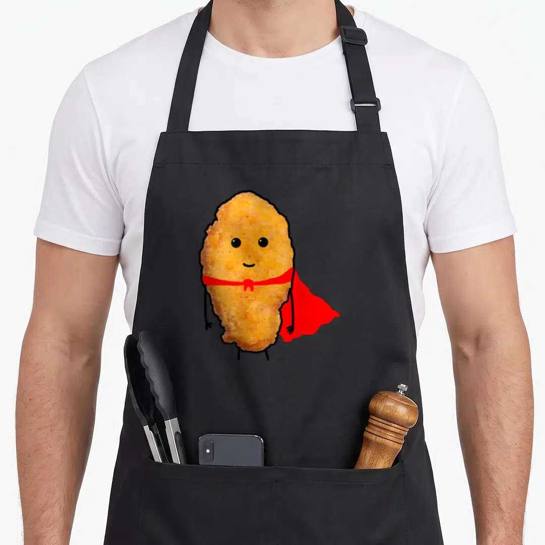 Hero chicken print apron - Cartoon golden brown chicken nugget wearing red cape with black limbs and smile — shown on a black cotton canvas apron against a white background