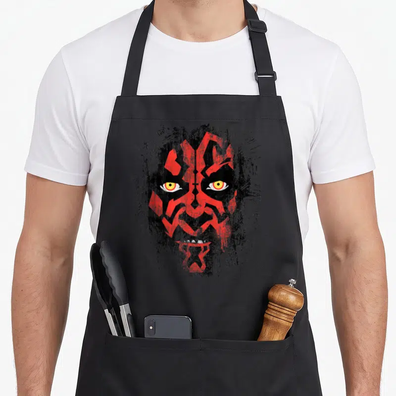 Weathered face apron - Distressed stylized face with red markings, black background, and yellow eyes — shown on a black cotton canvas apron against a white background