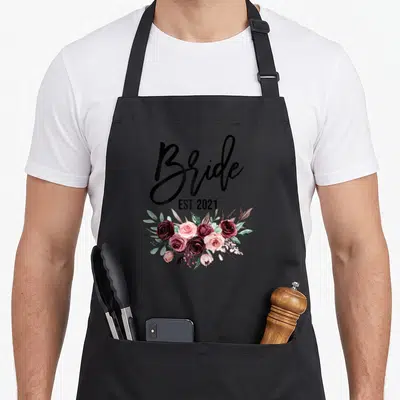 Est wedding day aprons - Bride EST. 2021 text above watercolor roses in burgundy and pink — shown on a black cotton canvas apron against a white background
