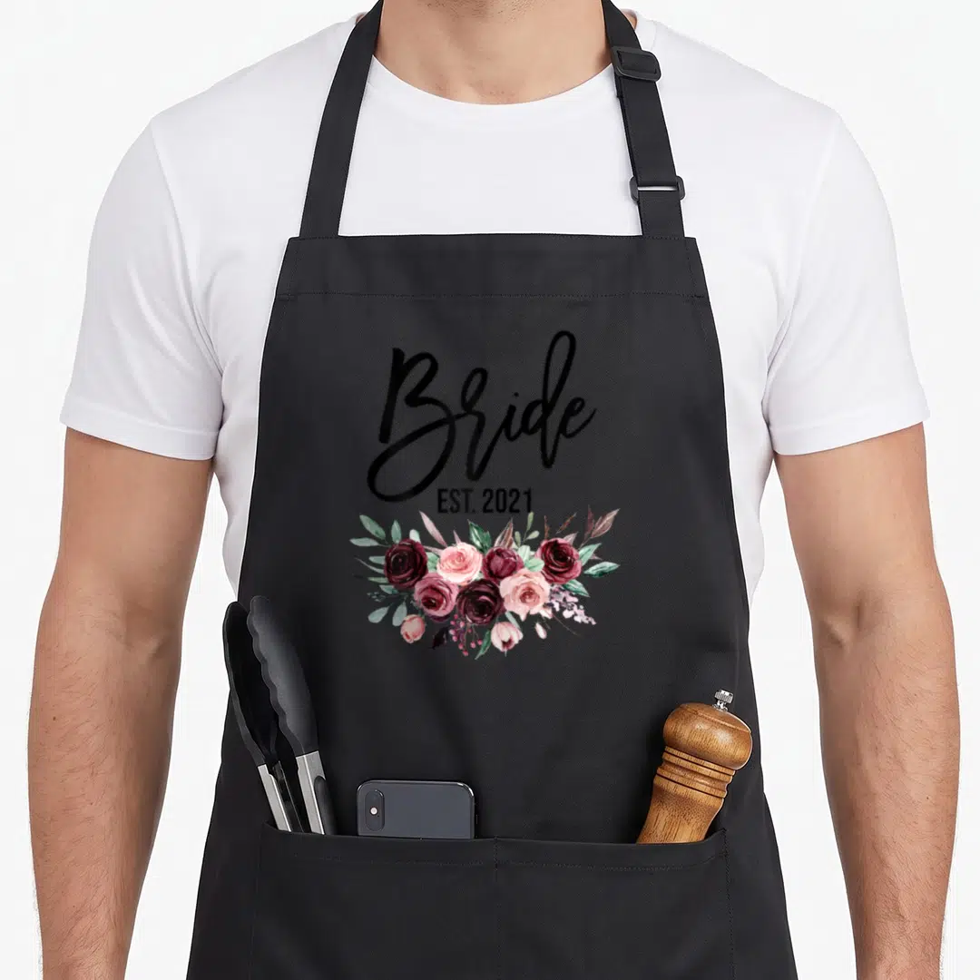Est wedding day aprons - Bride EST. 2021 text above watercolor roses in burgundy and pink — shown on a black cotton canvas apron against a white background