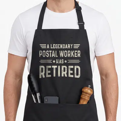 Retired legendary postal aprons - Beige distressed text reading A LEGENDARY POSTAL WORKER HAS RETIRED with stars — shown on a black cotton canvas apron against a white background