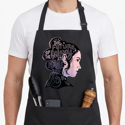 Rey nebula apron - Woman profile with THE FUTURE OF THE GALAXY IS FEMALE text in hair, stars, and symbol in black pink — shown on a black cotton canvas apron against a white background