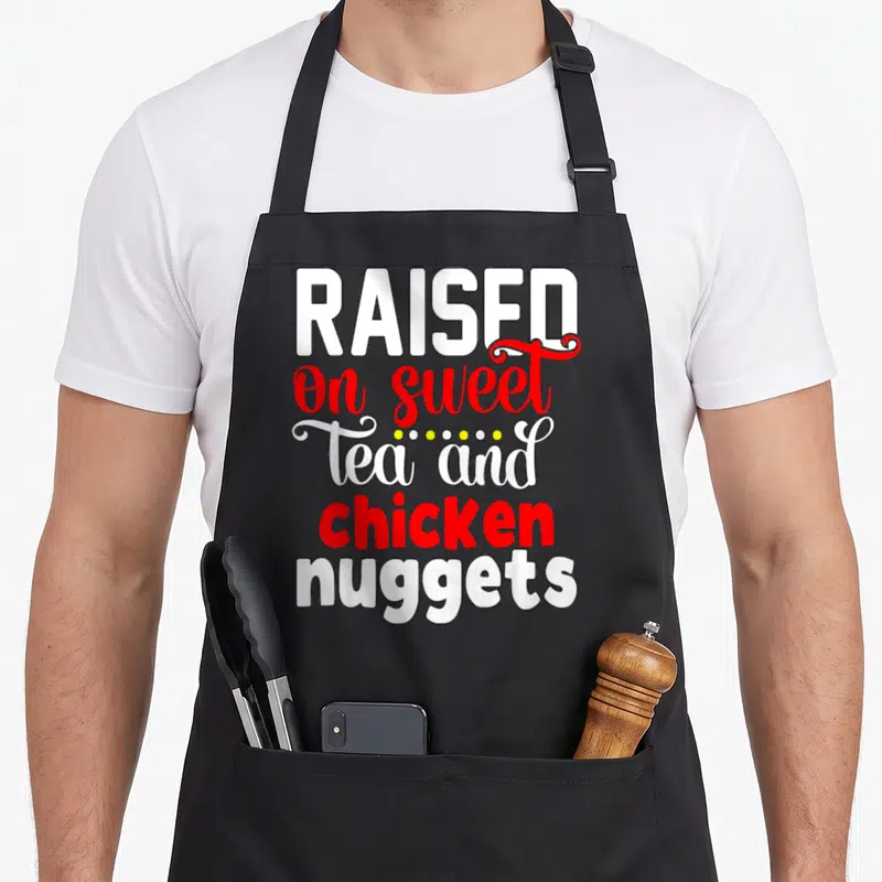 Raised chicken apron - Stacked white and red text reading raised on sweet tea and chicken nuggets — shown on a black cotton canvas apron against a white background