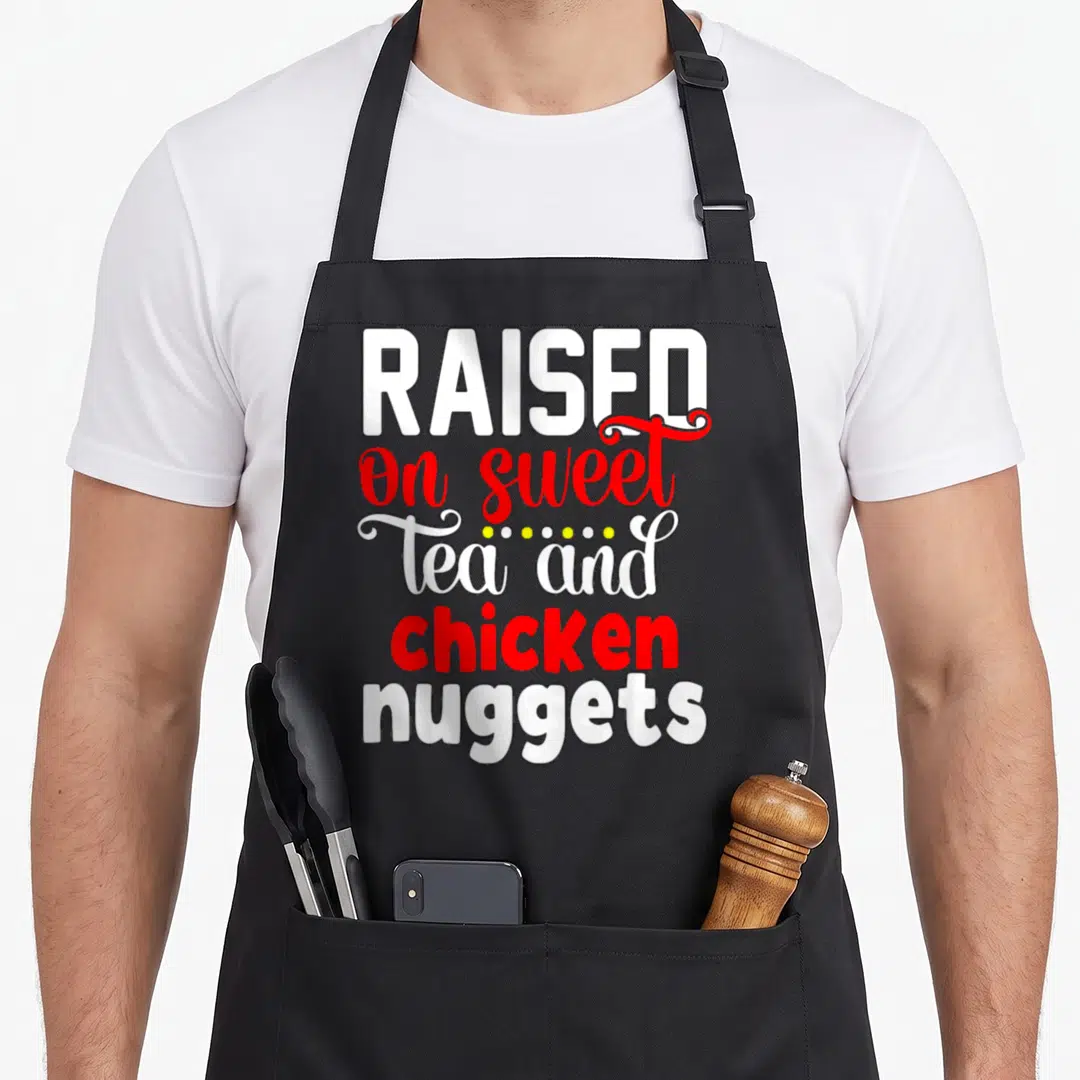 Raised chicken apron - Stacked white and red text reading raised on sweet tea and chicken nuggets — shown on a black cotton canvas apron against a white background