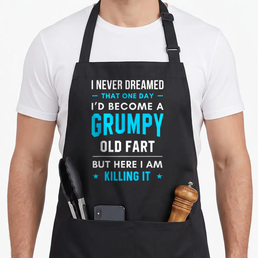 Dreamed old people gag for men apron - Stacked white and blue text reading I never dreamed become grumpy old fart — shown on a black cotton canvas apron against a white background
