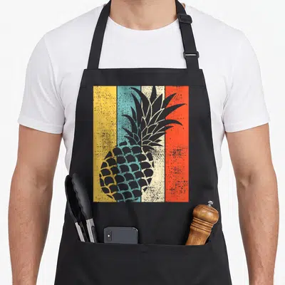 Summer pineapple apron - Grey pineapple silhouette over four vertical stripes in yellow, blue, white, and orange — shown on a black cotton canvas apron against a white background