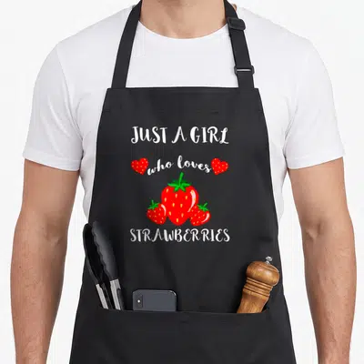 Funny fruit strawberry apron - White text Just A Girl who loves Strawberries with three red berries and two hearts — shown on a black cotton canvas apron against a white background