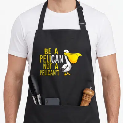 Alway pelican apron - Stacked yellow and white text reading Be a Pelican next to a grey cartoon pelican with a yellow — shown on a black cotton canvas apron against a white background