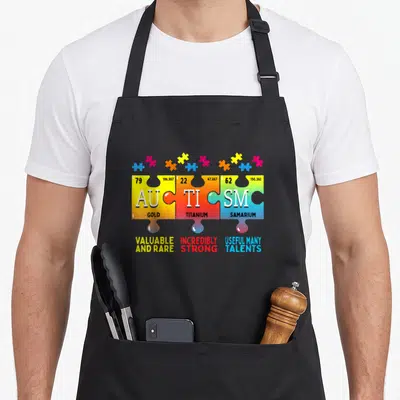 Autism inclusion awareness apron - Three colored puzzle pieces with chemical symbols spelling autism and descriptive — shown on a black cotton canvas apron against a white background