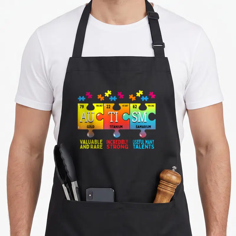 Autism inclusion awareness apron - Three colored puzzle pieces with chemical symbols spelling autism and descriptive — shown on a black cotton canvas apron against a white background