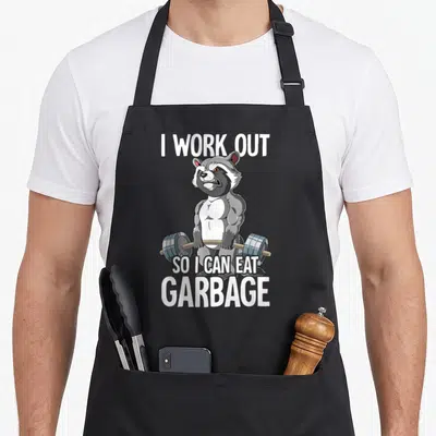 Raccoon training apron - Muscular cartoon raccoon holding a blue barbell with white text about eating garbage — shown on a black cotton canvas apron against a white background