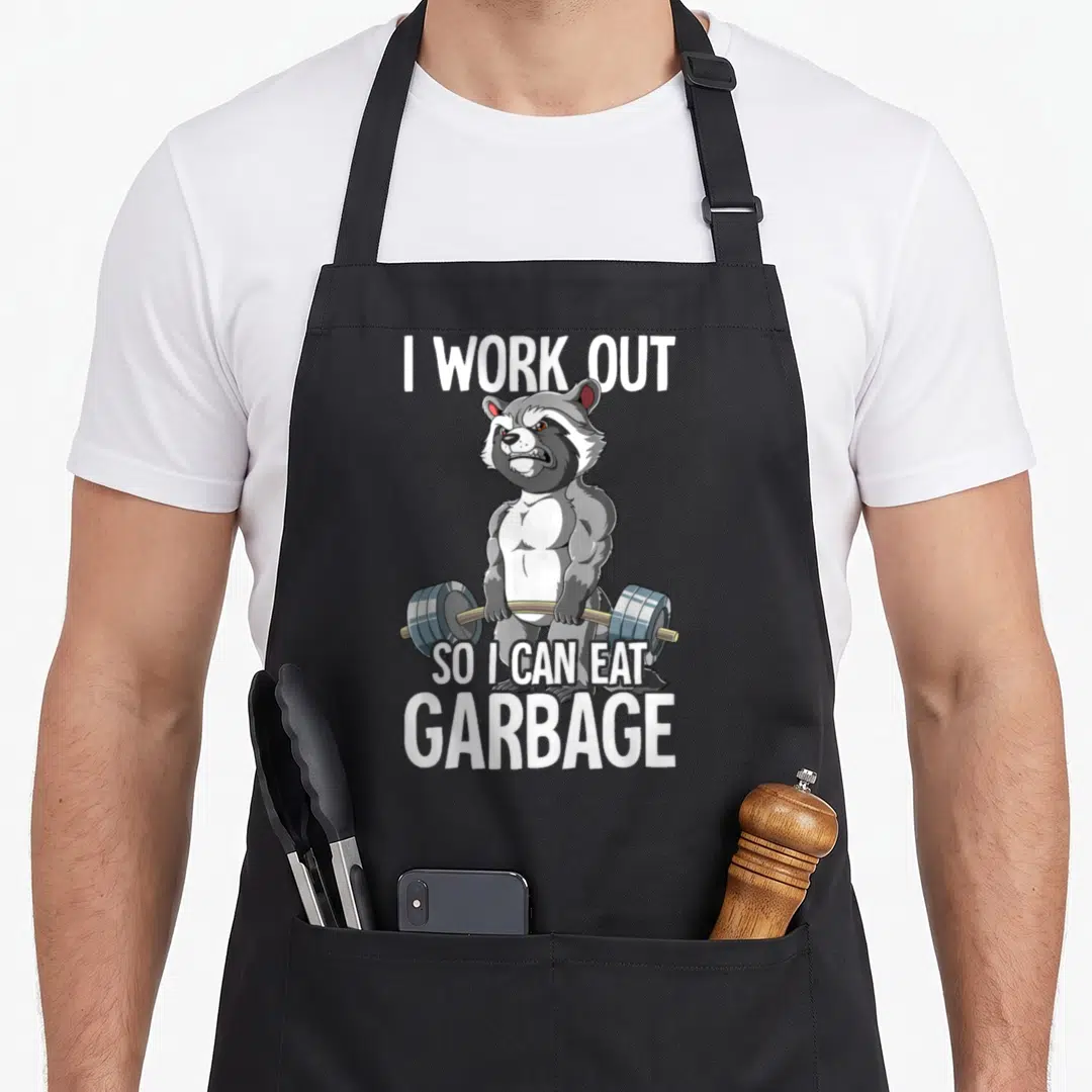 Raccoon training apron - Muscular cartoon raccoon holding a blue barbell with white text about eating garbage — shown on a black cotton canvas apron against a white background