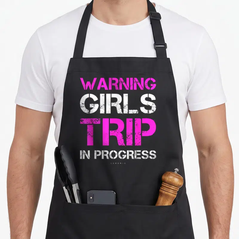 Warning girls trip apron - Stacked pink and white stenciled text reading warning girls trip in progress — shown on a black cotton canvas apron against a white background