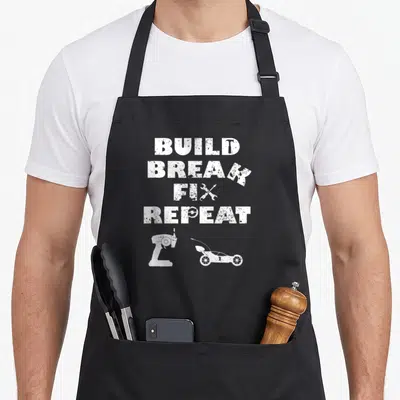 Fix car apron - White distressed BUILD BREAK FIX REPEAT text with hammer wrench bench grinder and go-kart silhouettes — shown on a black cotton canvas apron against a white background