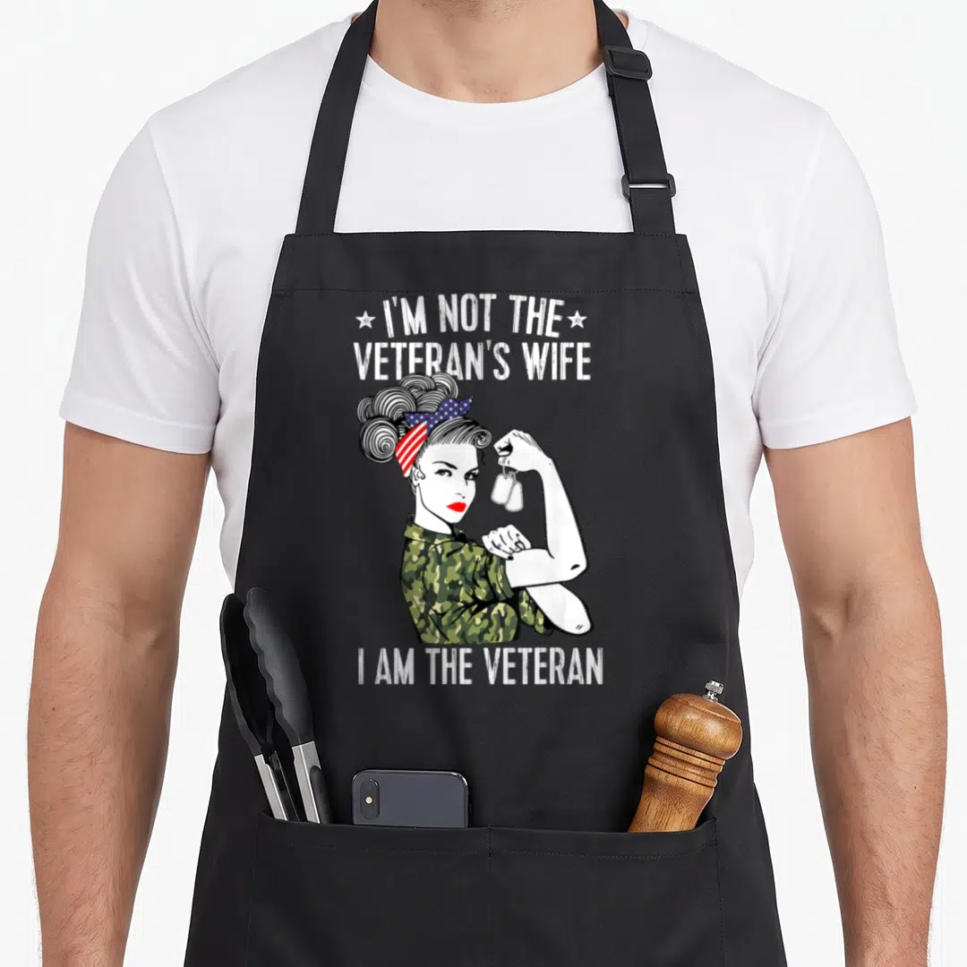 Wife patriotic apron - Cartoon woman in camo shirt and flag bandana flexing arm holding dog tags with text — shown on a black cotton canvas apron against a white background