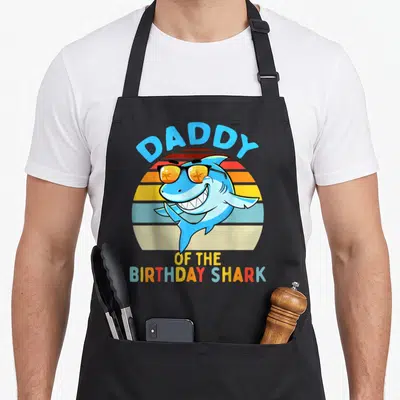 Shark family cooking aprons - Blue cartoon shark in sunglasses with retro sun and Daddy of the Birthday Shark text — shown on a black cotton canvas apron against a white background