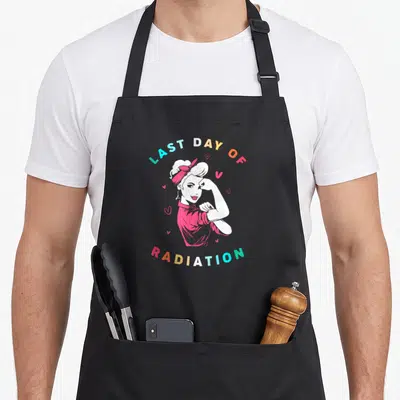 Strong radiation aprons - Cartoon woman flexing arm with Last Day of Radiation text and hearts — shown on a black cotton canvas apron against a white background