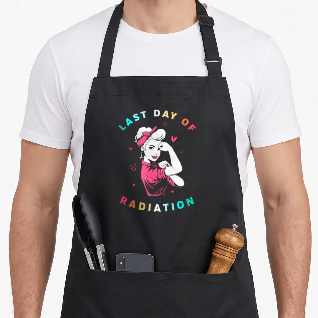 Strong radiation aprons - Cartoon woman flexing arm with Last Day of Radiation text and hearts — shown on a black cotton canvas apron against a white background