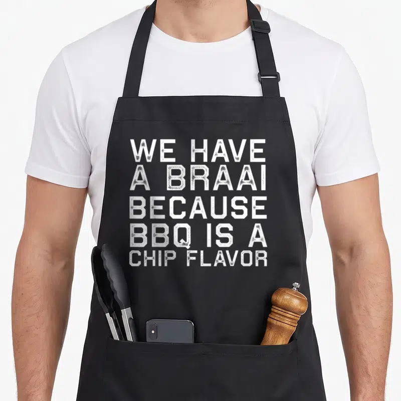 Braai family aprons - White stacked text reading We Have a Braai Because BBQ Is A Chip Flavor — shown on a black cotton canvas apron against a white background