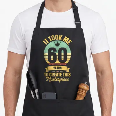 60th joke apron - Retro circular graphic with crown, number 60, and text IT TOOK ME 60 YEARS TO CREATE THIS Masterpiece — shown on a black cotton canvas apron against a white background