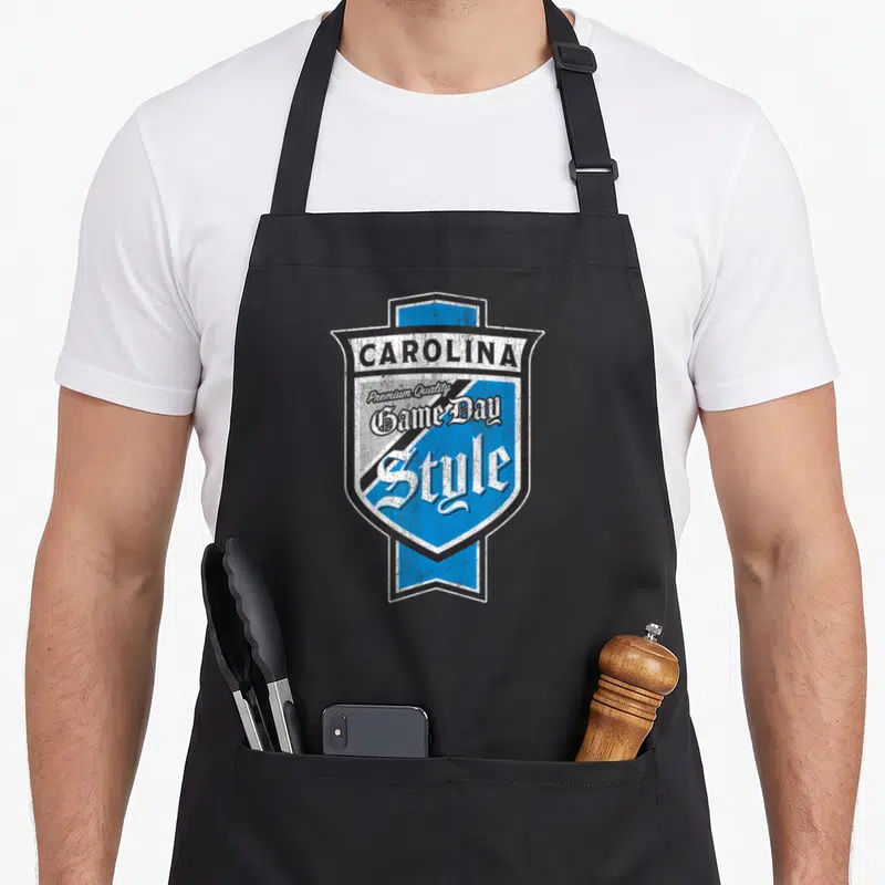 Faded beer apron - Retro shield with stacked text CAROLINA Premium Quality Game Day Style in blue white and black — shown on a black cotton canvas apron against a white background