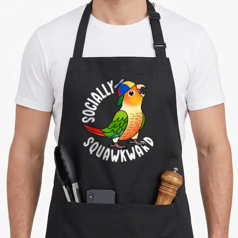 Socially pineapple apron - Cartoon parrot in colorful cap with white arched text reading SOCIALLY SQUAWKWARD — shown on a black cotton canvas apron against a white background