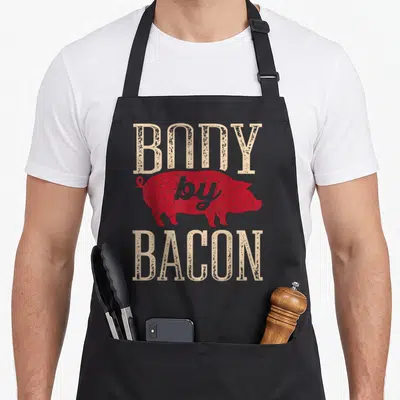 Bacon food aprons - Red pig silhouette with distressed beige text reading Body by Bacon — shown on a black cotton canvas apron against a white background