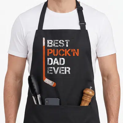Hockey puckn father day apron - Orange hockey stick with BEST PUCKIN DAD EVER distressed text — shown on a black cotton canvas apron against a white background
