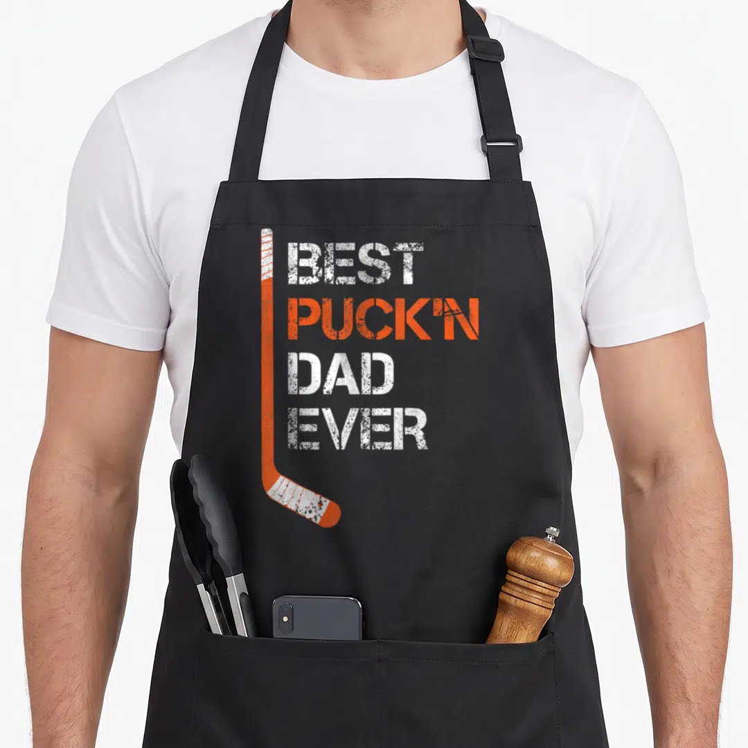 Hockey puckn father day apron - Orange hockey stick with BEST PUCKIN DAD EVER distressed text — shown on a black cotton canvas apron against a white background