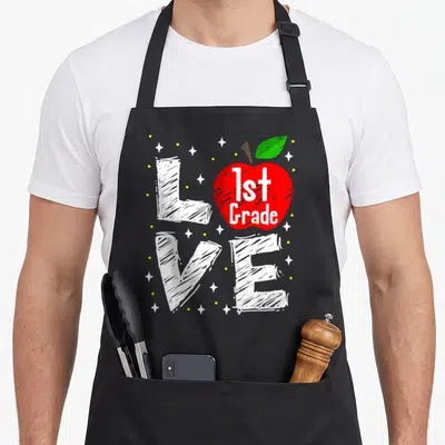 Apple art teacher aprons - Red apple with 1st Grade text inside sketched white LOVE letters surrounded by stars — shown on a black cotton canvas apron against a white background