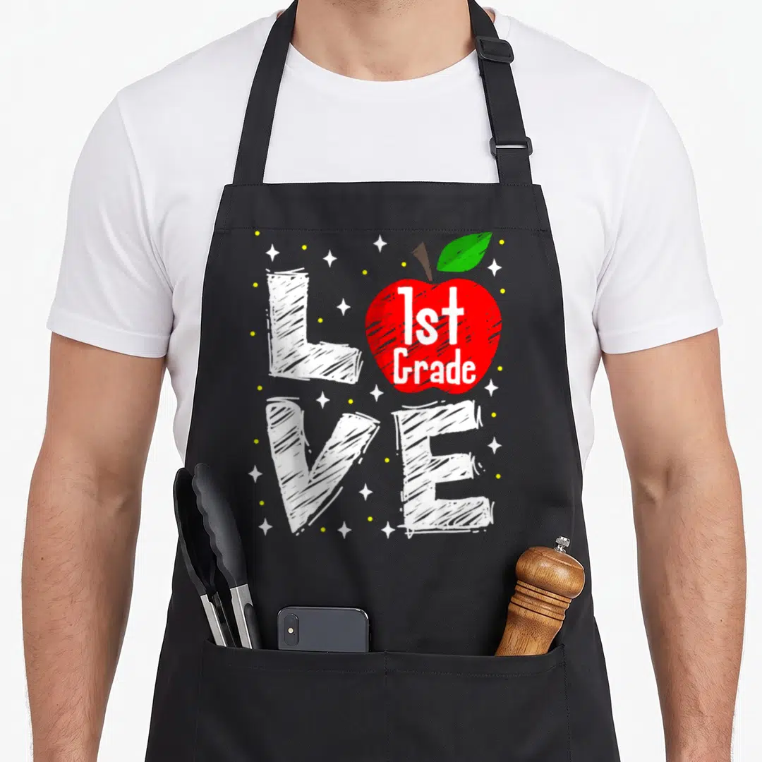 Apple art teacher aprons - Red apple with 1st Grade text inside sketched white LOVE letters surrounded by stars — shown on a black cotton canvas apron against a white background