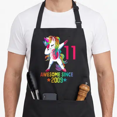 11th unicorn birthday apron - Dabbing white unicorn with rainbow mane next to pink 11 and AWESOME SINCE 2009 text — shown on a black cotton canvas apron against a white background