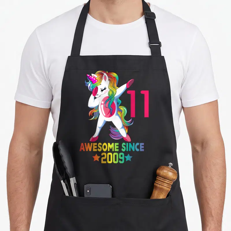 11th unicorn birthday apron - Dabbing white unicorn with rainbow mane next to pink 11 and AWESOME SINCE 2009 text — shown on a black cotton canvas apron against a white background