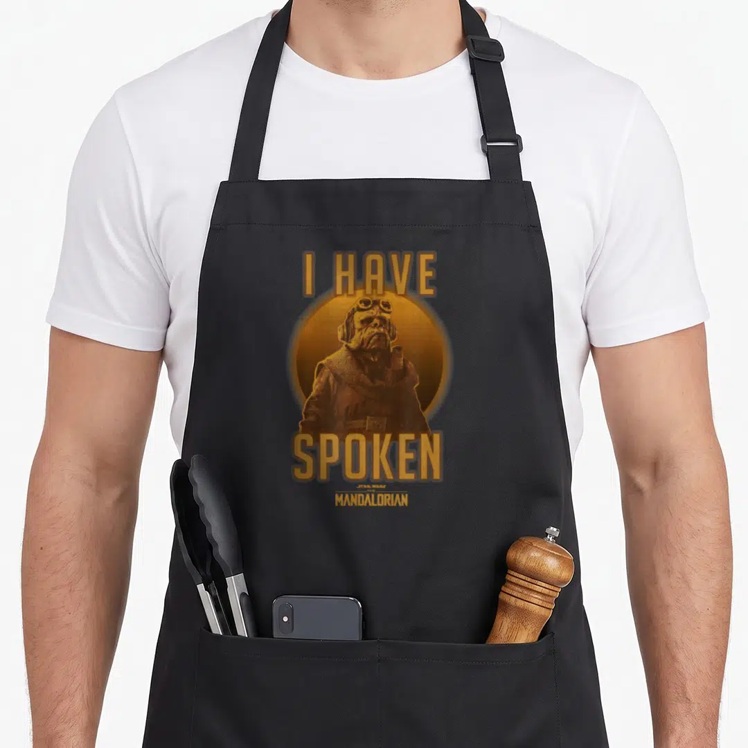 Kuiil de apron - Kuiil character in gold circle with I Have Spoken text above and below — shown on a black cotton canvas apron against a white background