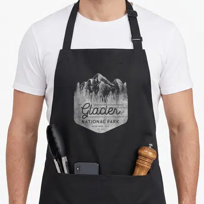 Glacier family aprons - White distressed mountain illustration with Glacier National Park Montana USA text — shown on a black cotton canvas apron against a white background