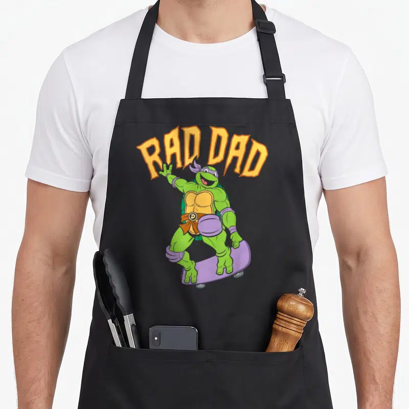 Skateboard father day apron - Arched RAD DAD text above a green turtle standing on a purple skateboard wearing pads — shown on a black cotton canvas apron against a white background