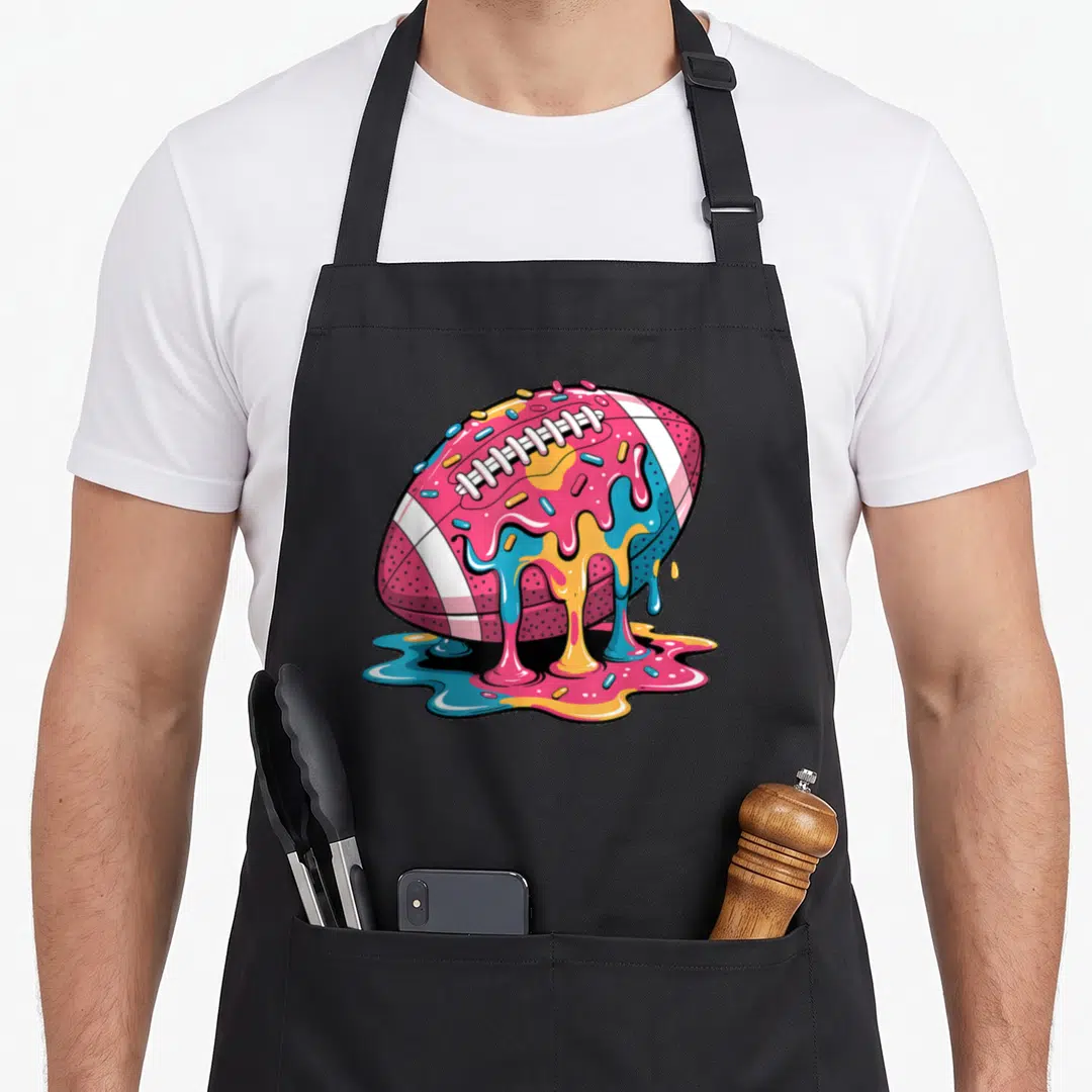 Football drip apron - Cartoon football shaped like a donut with pink blue and yellow dripping glaze and sprinkles — shown on a black cotton canvas apron against a white background